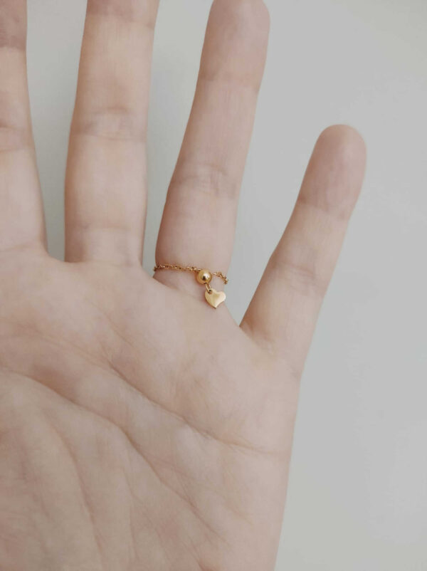 Anel ajustável (até ao tamanho 18) em aço com fio fino, retângulo com pedra vermelha à frente e travão com pequeno coração atrás, dourado